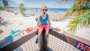 A child sitting on a balcony overlooking a beach with boats in the water, a sign reading "TOAST & PLAT DU JOUR," and a view of the city in the background. - Practically Perfect Place in Every Way (Pensacola)