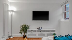 A modern living room with a flat-screen TV mounted on the wall, a white entertainment unit below, a potted plant, and a round coffee table. - 2 Bedroom Service Apt in Artillery Lane (London)