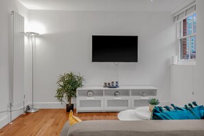 A modern living room with a flat-screen TV mounted on the wall, a white entertainment unit below, a potted plant, and a round coffee table. - 2 Bedroom Service Apt in Artillery Lane (London)