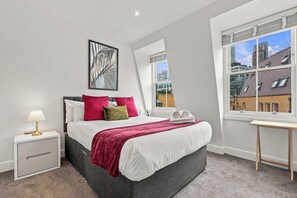 A modern bedroom with a bed, bedside tables, a lamp, a window with a view of buildings, and a framed picture on the wall. - 2 Bedroom Service Apt in Artillery Lane (London)