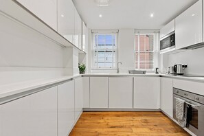 A modern kitchen with white cabinetry, a wooden floor, a window with blinds, and a built-in oven. - 2 Bedroom Service Apt in Artillery Lane (London)