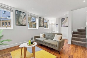 A modern living room with a grey sofa, a small round table with candles, and a staircase to the right. - 2 Bedroom Service Apt in Artillery Lane (London)