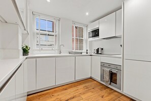 A modern kitchen with white cabinetry, a wooden floor, and a window with blinds. - 2 Bedroom Service Apt in Artillery Lane (London)