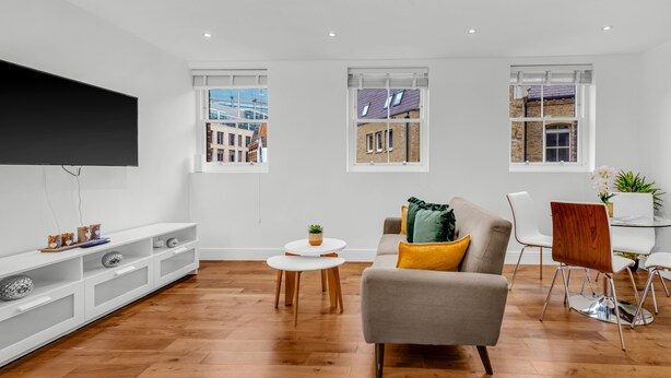A modern living room with a grey sofa, white chairs, a TV, and a dining table. - 2 Bedroom Service Apt in Artillery Lane (London)