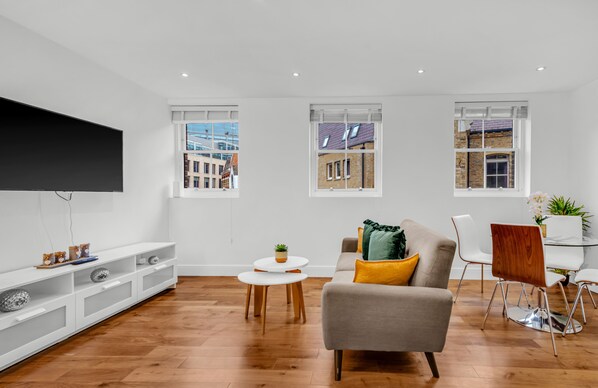 A modern living room with a grey sofa, white chairs, a TV, and a dining table. - 2 Bedroom Service Apt in Artillery Lane (London)