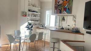 Un espace de vie moderne comprenant un canapé gris, une table à manger en verre avec des chaises, et un coin cuisine doté d’un plan de travail en bois et d’étagères.
