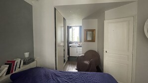 Une chambre à coucher avec un lit, une table de chevet avec des livres, une chaise, une porte et une salle de bain visible au fond.