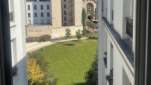 Une vue depuis une fenêtre donnant sur une cour intérieure, avec des bâtiments, dont une église dotée d’un clocher, et un ciel bleu dégagé.