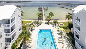 A swimming pool surrounded by a white building with balconies, a dock extending into the water, and palm trees.