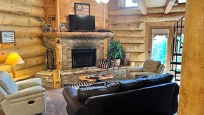 Ein Blockhauss Inneres mit Kamin aus Stein, einer Ledersofa und einem Fernseher auf einem Regal.
