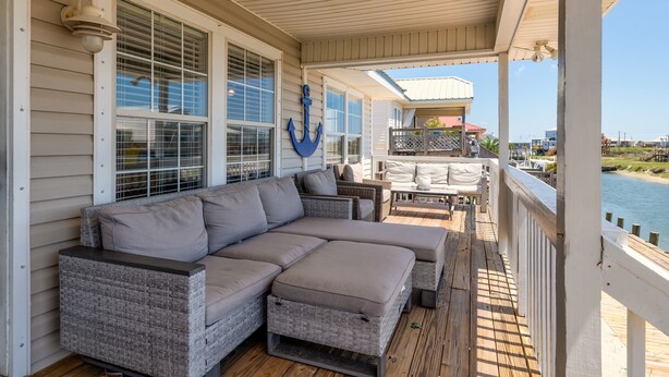 A covered porch with a wicker sectional sofa, a dining table with chairs, and a view of the water.