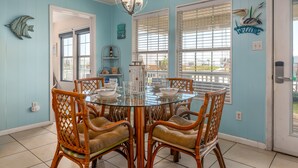 A dining area with a round glass table, wicker chairs, and a view through large windows.