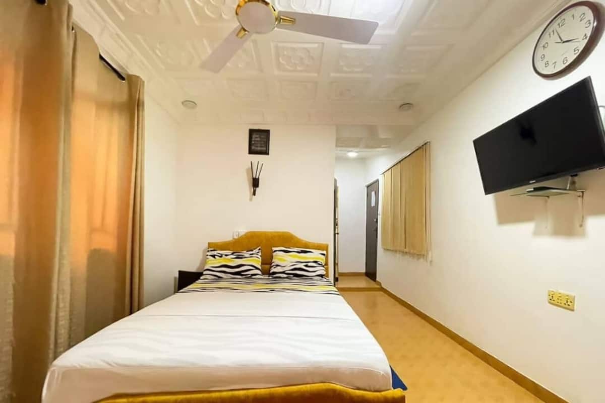 A hotel room with a bed, a ceiling fan, a clock, and a TV mounted on the wall.