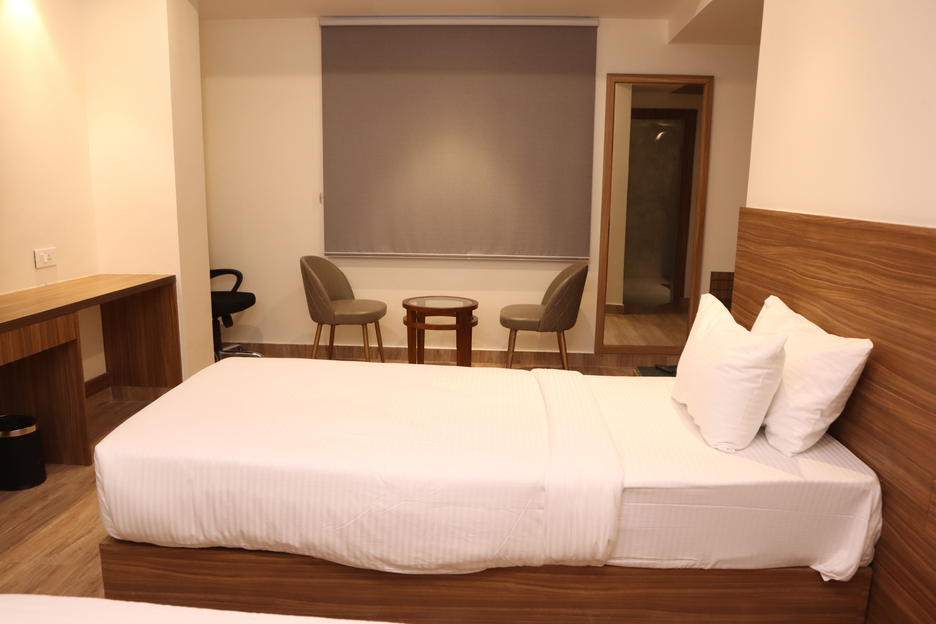 A hotel room with a large bed, a small table, two chairs, a large window with a view, and a mirror.
