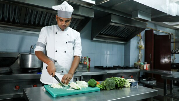 A chef in a professional kitchen with stainless steel surfaces, a large cutting board, and various vegetables. - Hotel Sharda Residency (Patna)