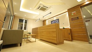 A modern reception area with a wooden reception desk, a seating area, and a large window. - Hotel Sharda Residency (Patna)
