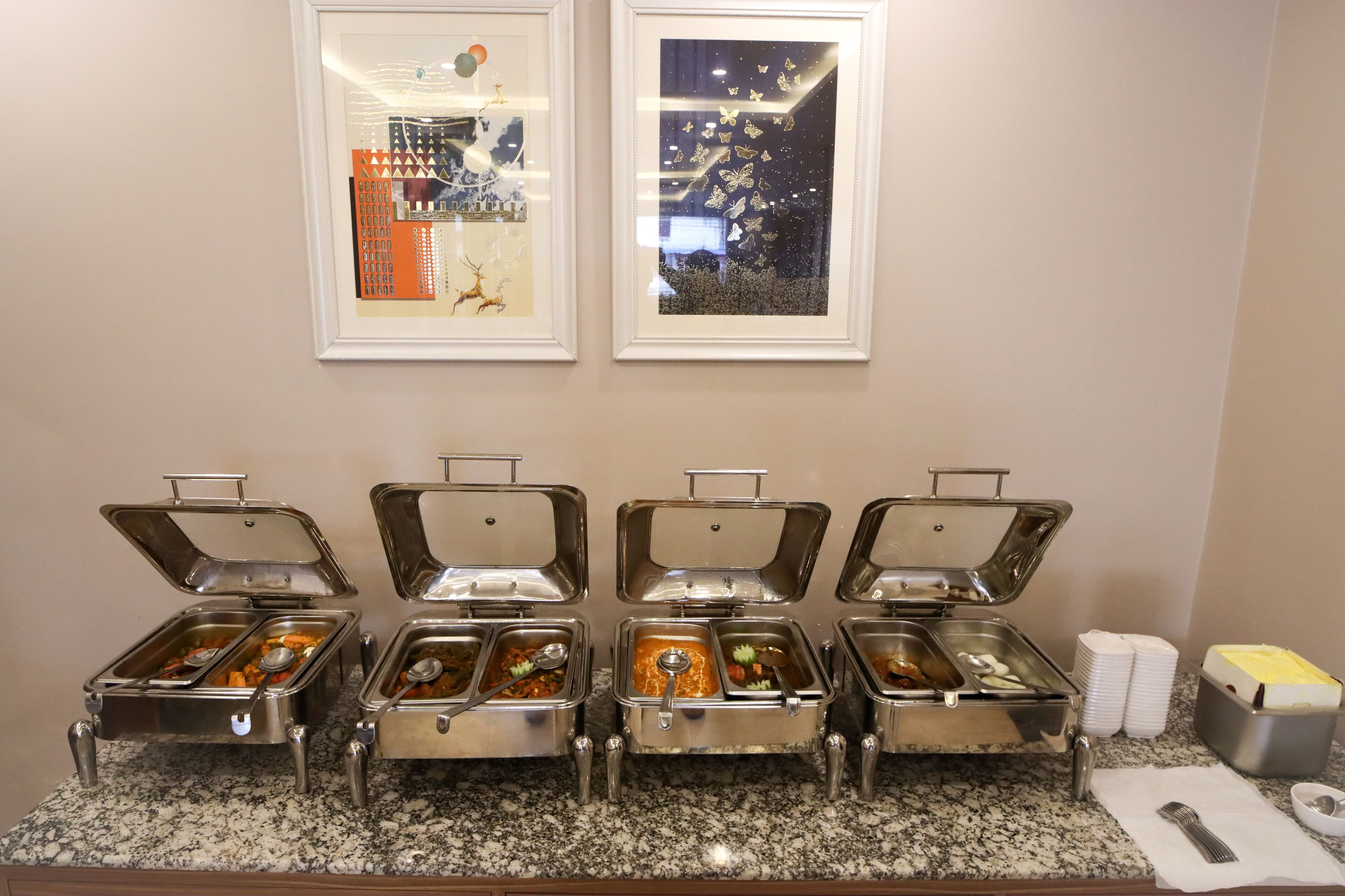 A buffet setup with multiple food dispensers on a granite countertop, framed pictures on the wall, and a napkin holder with utensils.