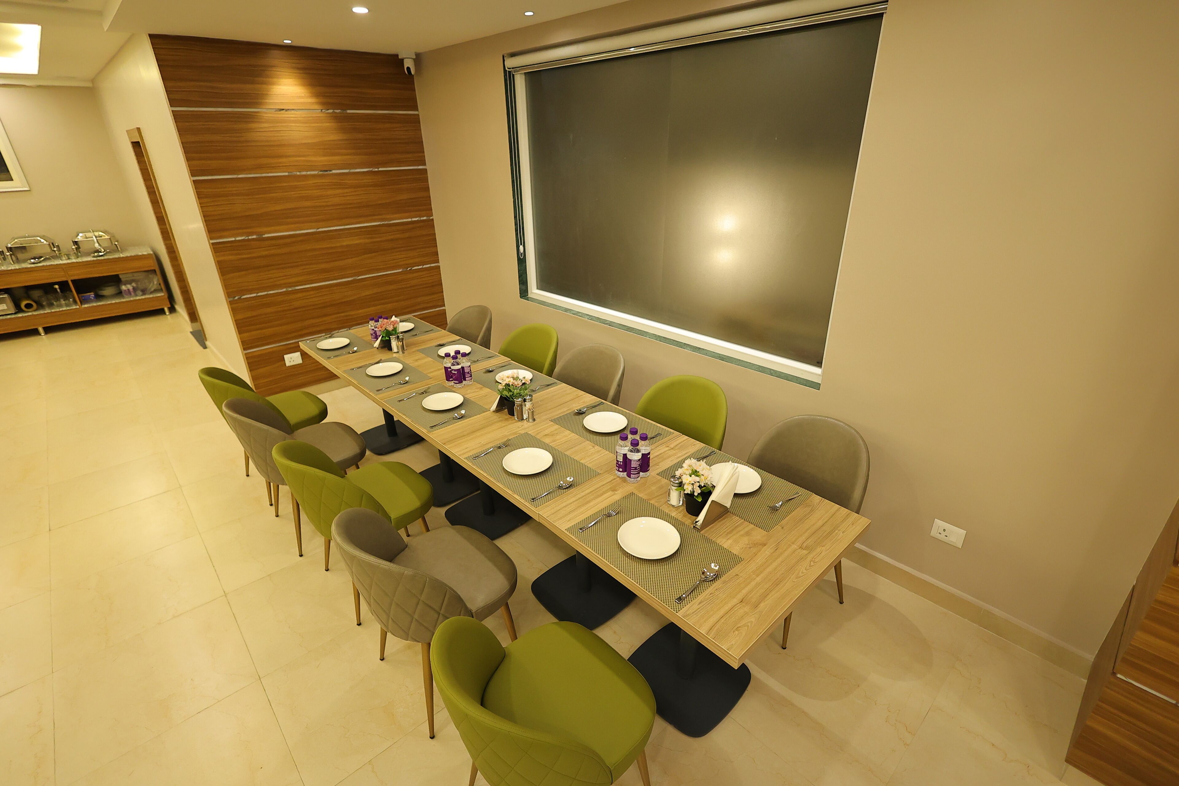 A modern dining area with a long table set for six, surrounded by green and gray chairs. A wooden wall and a large window are visible in the background.