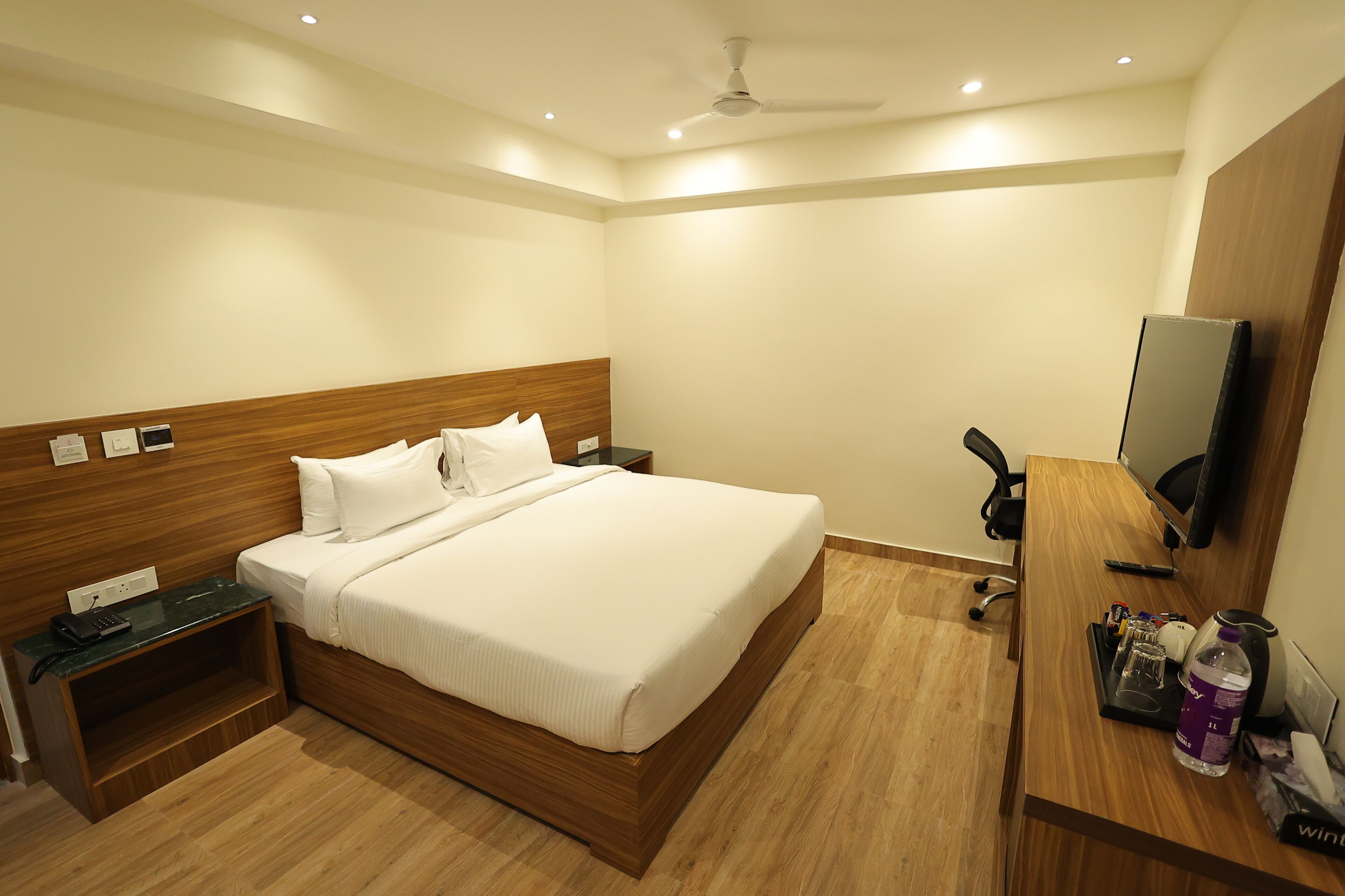 A hotel room with a wooden headboard, a bed with white linens, a desk with a computer, a chair, and a television on the wall.