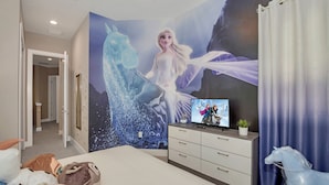 Un dormitorio con un mural grande de un personaje montando un caballo, un televisor sobre un aparador, y una cama con almohadas y peluches.