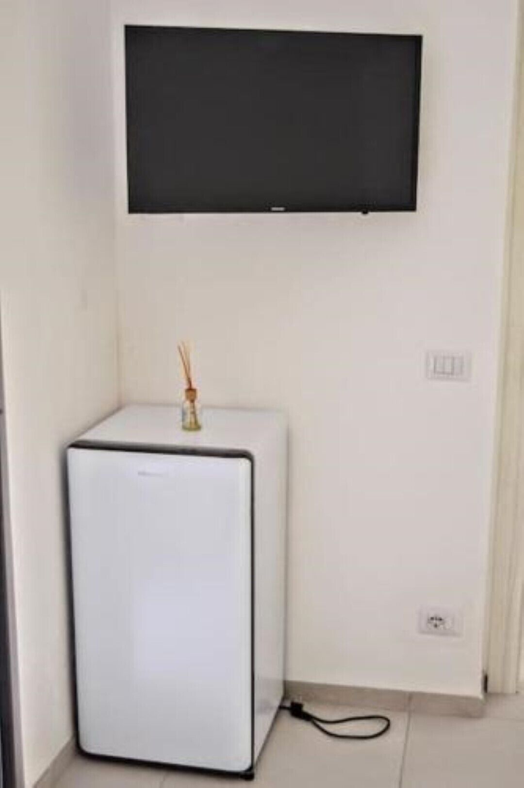 A small white refrigerator with a black top, a wall-mounted TV above it, and a power outlet on the wall.