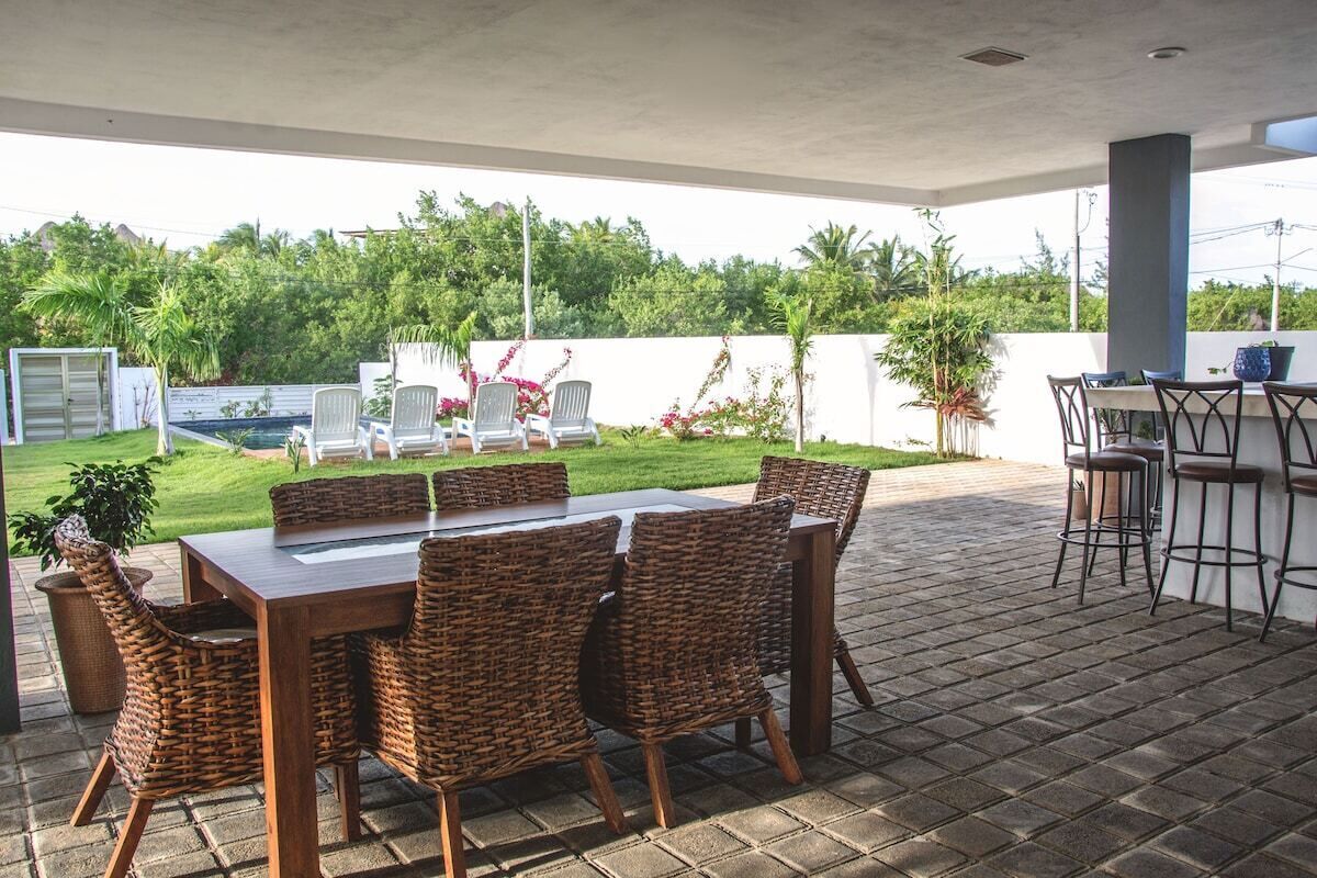 An outdoor patio area with a wooden table and wicker chairs, a bar area with stools, and a view of a lush garden and pool.