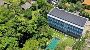 Vista aérea de un hotel con piscina, rodeado de abundante vegetación y un edificio grande con varios balcones.