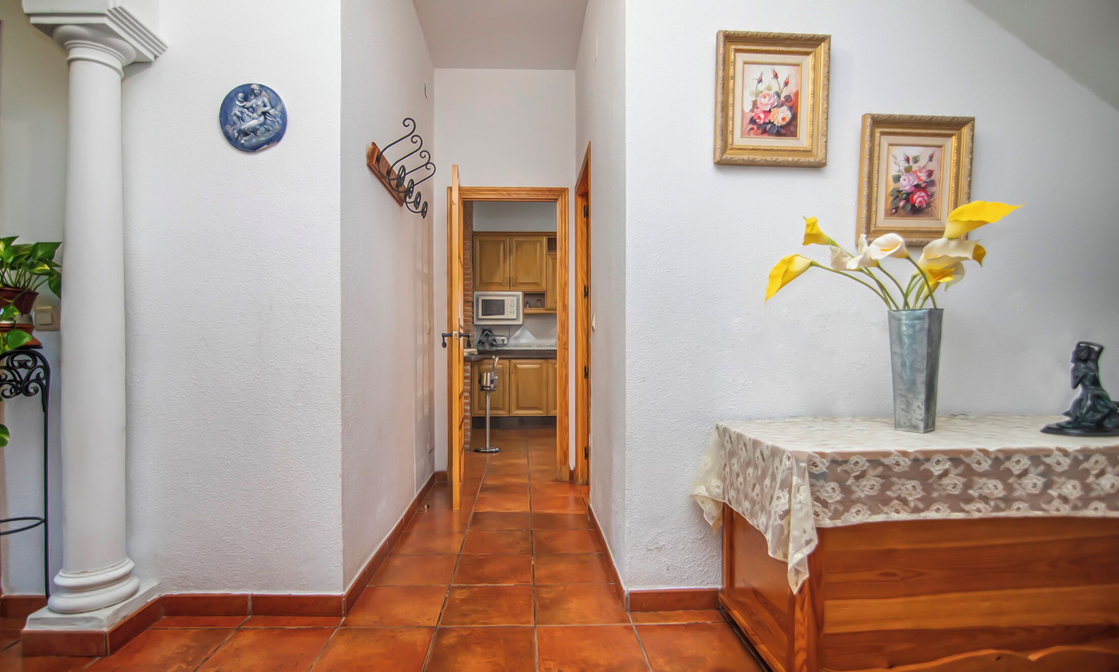 Un pasillo estrecho con suelo de baldosas, una puerta de madera que da a una cocina y una pequeña mesa con un jarrón de flores.