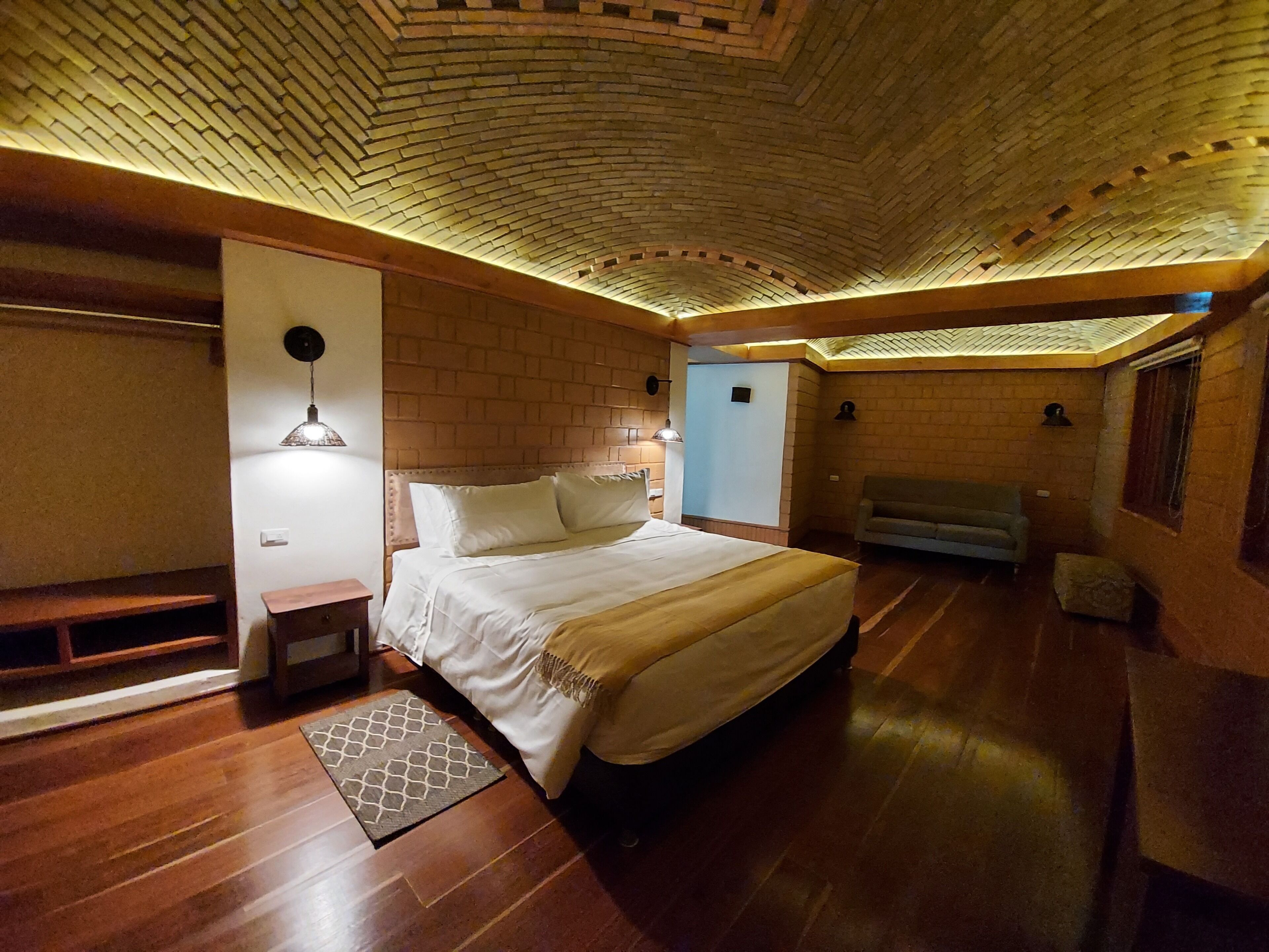 A room with a brick-patterned ceiling, a bed with white bedding, a bench, a sofa, and a patterned rug.