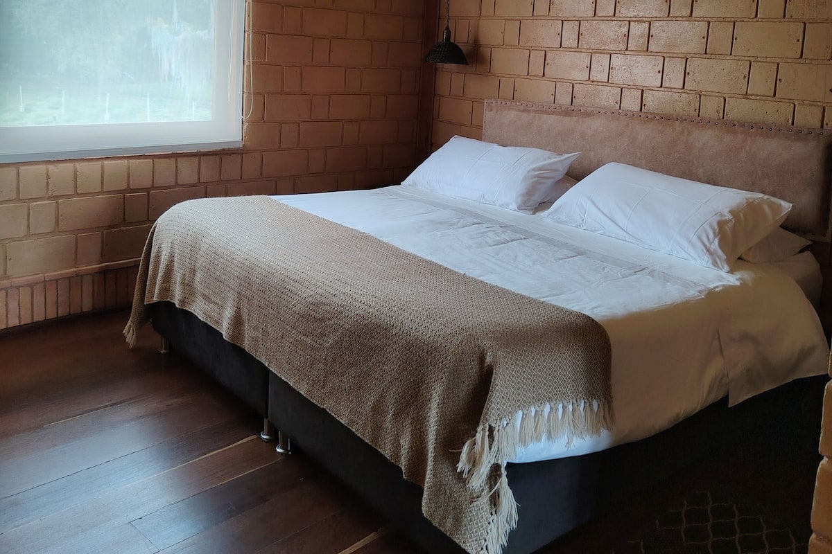 A bedroom with a brick-patterned ceiling, a large window, a bed with white linens, and a wooden floor.