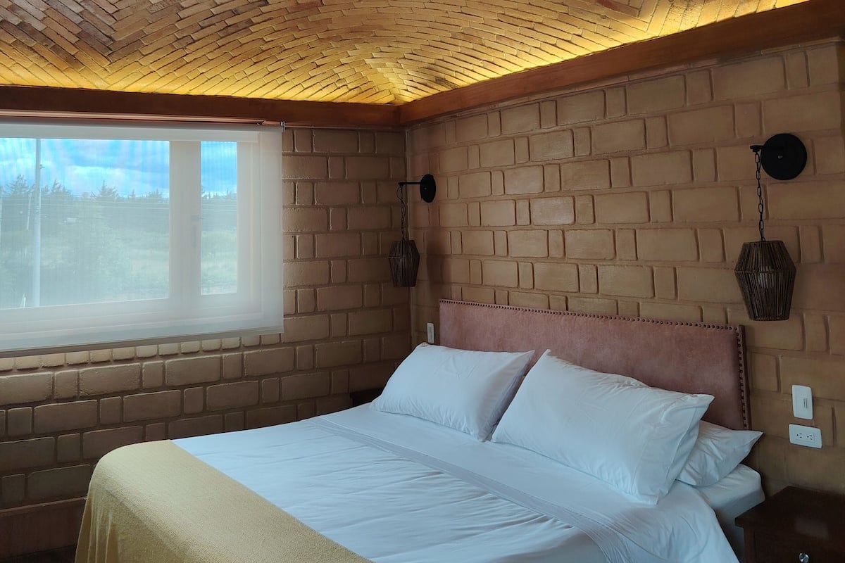 A room with a brick-patterned ceiling, a bed with white bedding, a window, and a wall-mounted lamp.