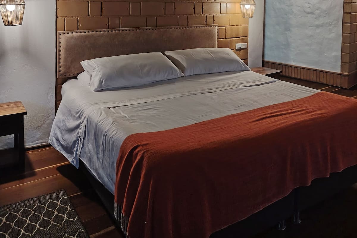A bedroom with a brick-patterned wall, a tufted headboard, a bed with white linens, a red throw blanket, and two wall-mounted lamps.