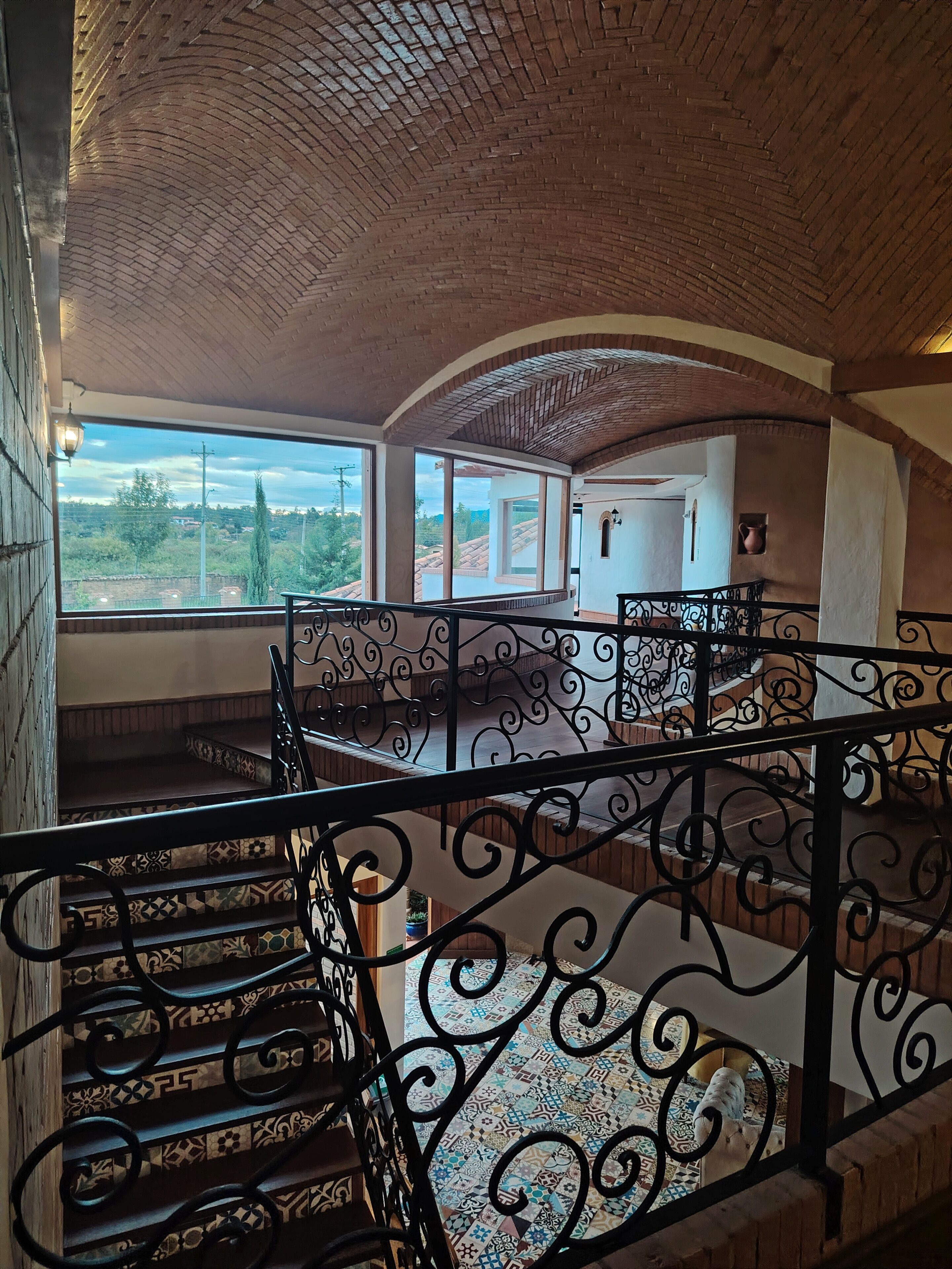 Una escalera con barandillas negras ornamentales, baldosas de suelo con diseños y vistas al exterior con vegetación.