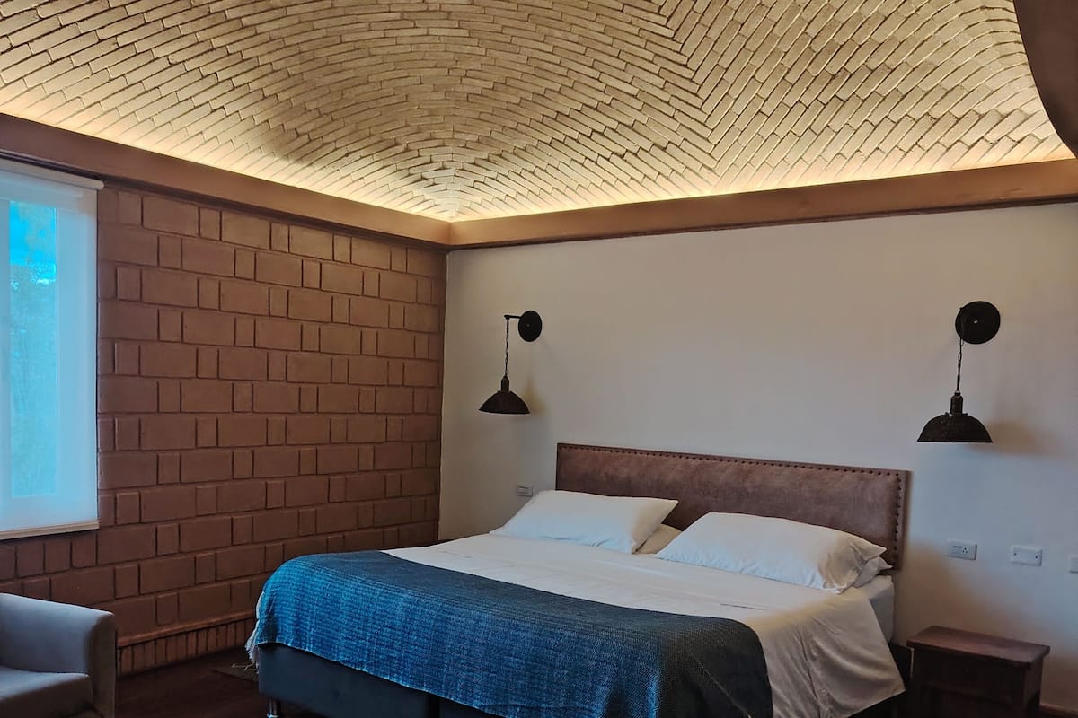 A bedroom with a brick wall, a wooden floor, a bed with a blue blanket, two bedside lamps, and a small wooden nightstand.