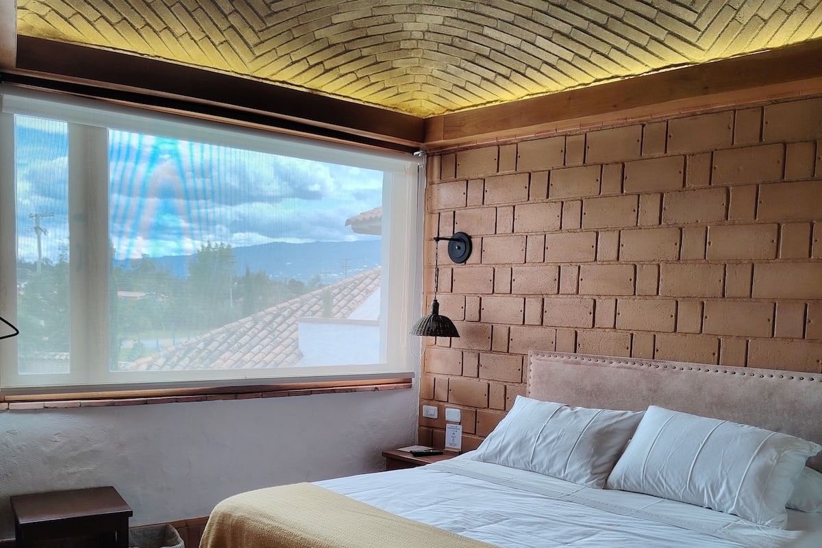 A bedroom with a brick-patterned headboard, a large bed with white linens, a bedside table, and a window with a view of the outside.