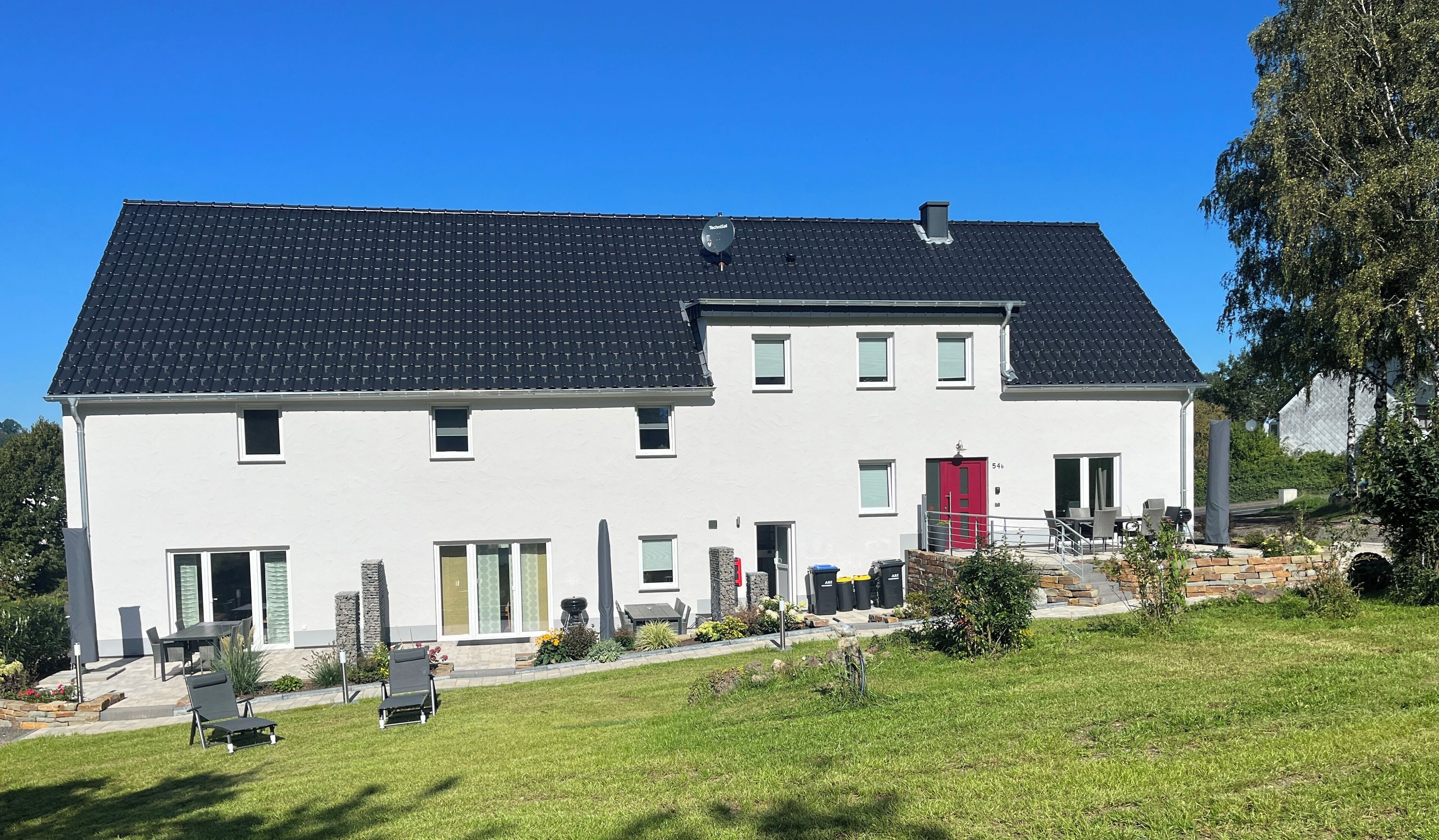Une maison à deux étages, avec un toit recouvert de tuiles sombres, des murs extérieurs blancs et une porte rouge. Un petit jardin, avec une pelouse et quelques plantes en pot, se trouve devant la maison.