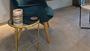 Ein modernes Wohnzimmer mit einem türkisfarbenen Sessel, einem runden Tisch mit Kerze, einem Flachbildfernseher und einer Wanduhr.
