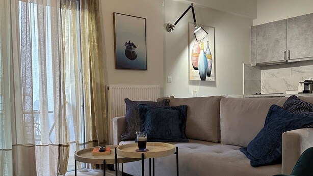 Ein modernes Wohnzimmer mit einer Couch, einem Couchtisch und einem Sessel. Ein Fenster mit durchsichtigen Vorhängen, ein Bild an der Wand und eine Küchenzeile im Hintergrund.