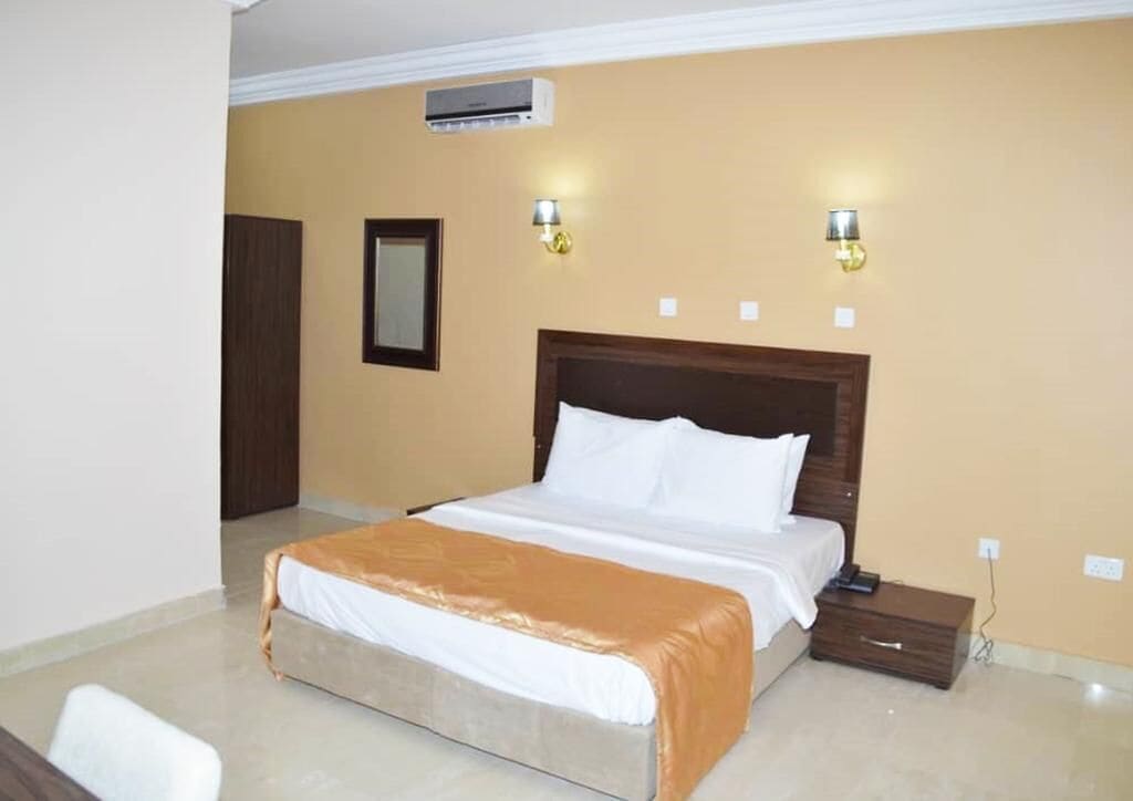 A hotel room with a large bed, a nightstand, two wall-mounted lamps, and an air conditioning unit.
