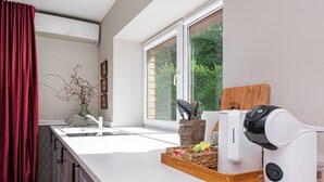 Eine moderne Küche mit weißer Arbeitsplatte, einer Kaffeemaschine, einem Fenster mit Blick auf Grünflächen und einem roten Vorhang.