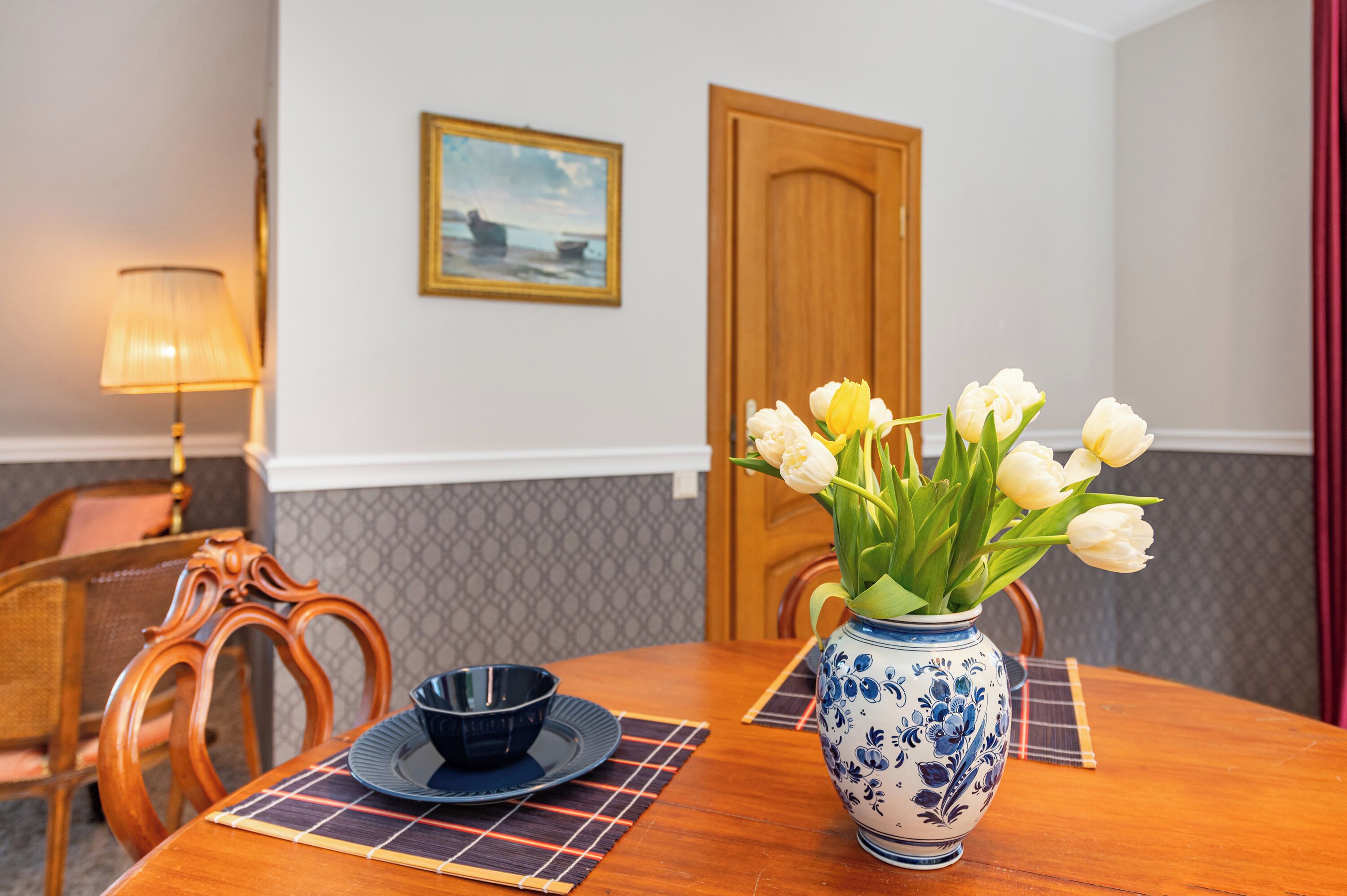 A dining table with a vase of white tulips, a blue and white ceramic vase, a black bowl, and a placemat.