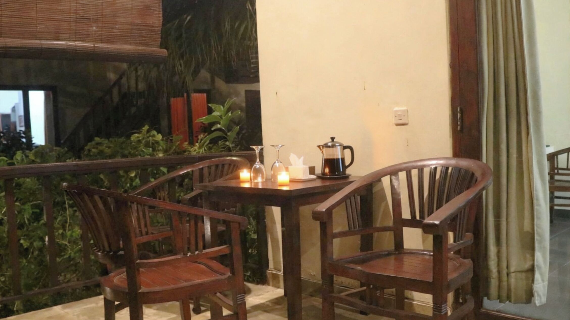 Uma varanda com duas cadeiras de madeira, uma mesa com cafeteira, velas e um vaso.