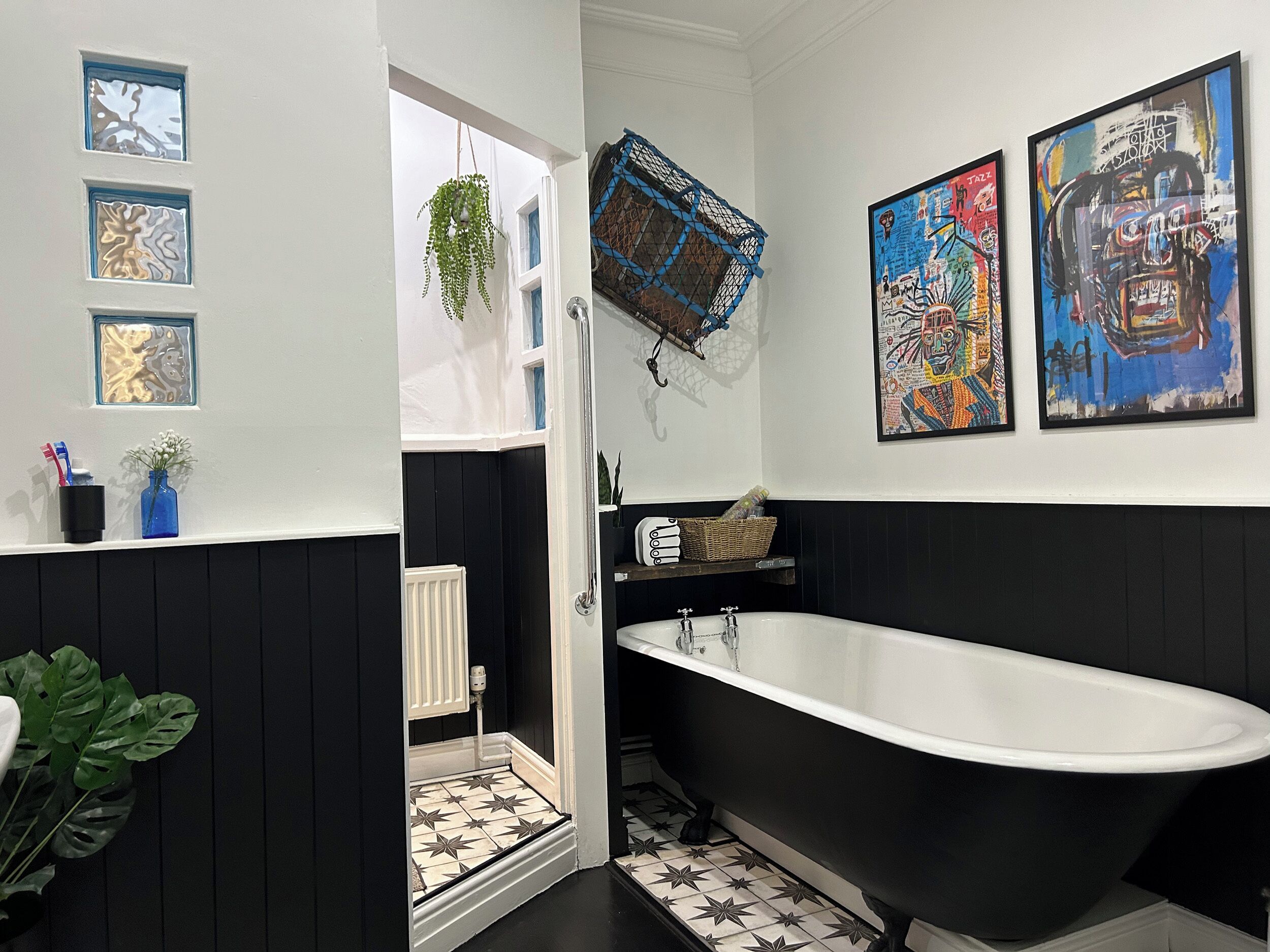 A bathroom with a black bathtub, white tiles, and black paneling. There are framed artworks on the wall and a hanging plant.