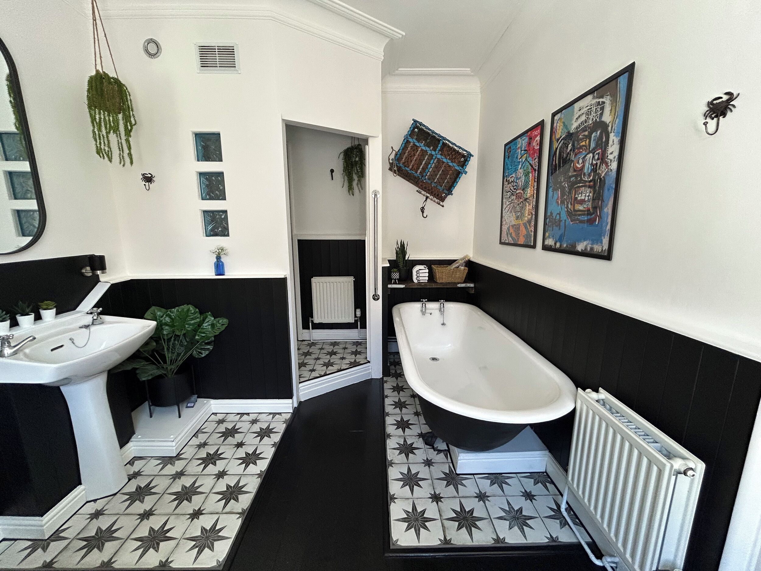 A bathroom with a black and white tiled floor, a white sink, a black bathtub, and a white radiator. There are framed pictures on the wall and a hanging plant.