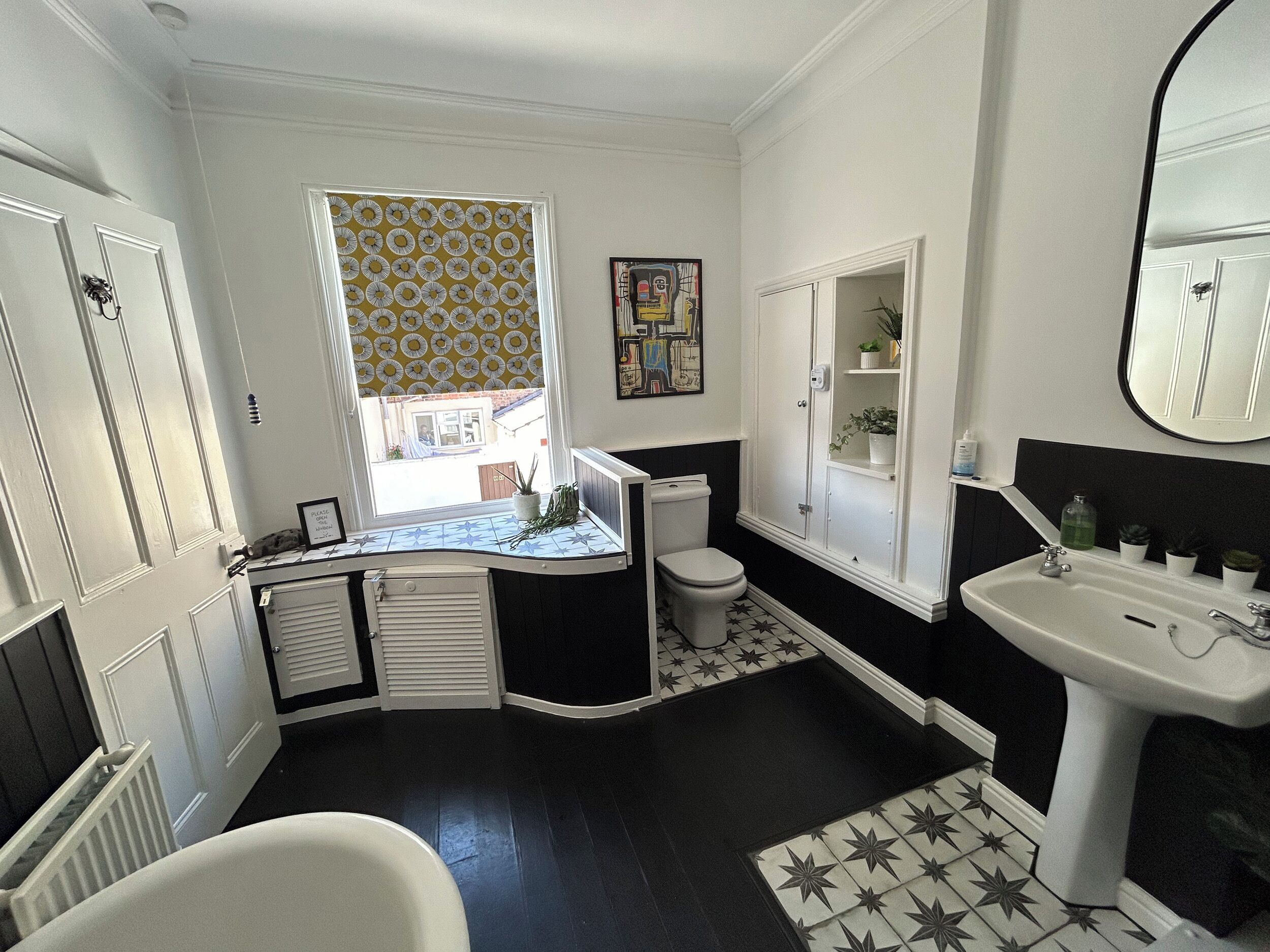 Une salle de bain avec une baignoire blanche, un lavabo blanc, des toilettes et une fenêtre ornée d’un rideau à motifs jaunes.