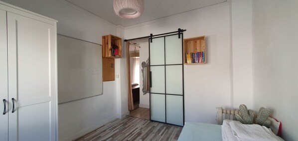 A bedroom with a sliding door, a bed with a white cover, a radiator, and a shelf with books.
