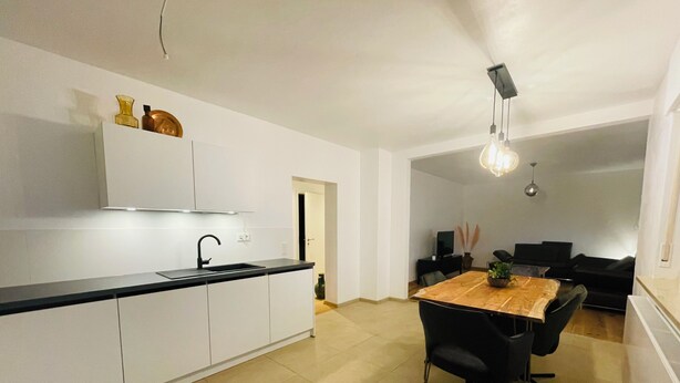 Une cuisine moderne avec des meubles blancs, un plan de travail noir et un coin repas comprenant une table en bois et des chaises noires.