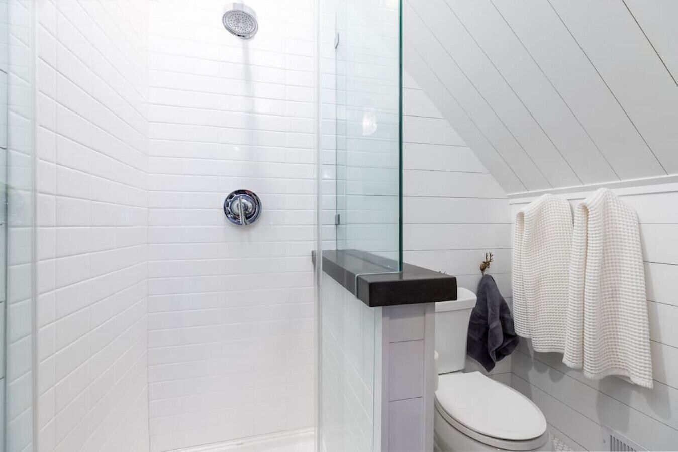 A modern bathroom with white tiled walls, a glass shower enclosure, a black countertop, and a toilet with a towel rack.