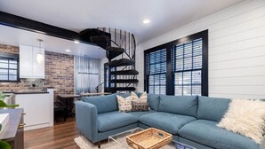 Un salon moderne avec un canapé d’angle bleu, une cuisine avec un mur en briques et un escalier en colimaçon.
