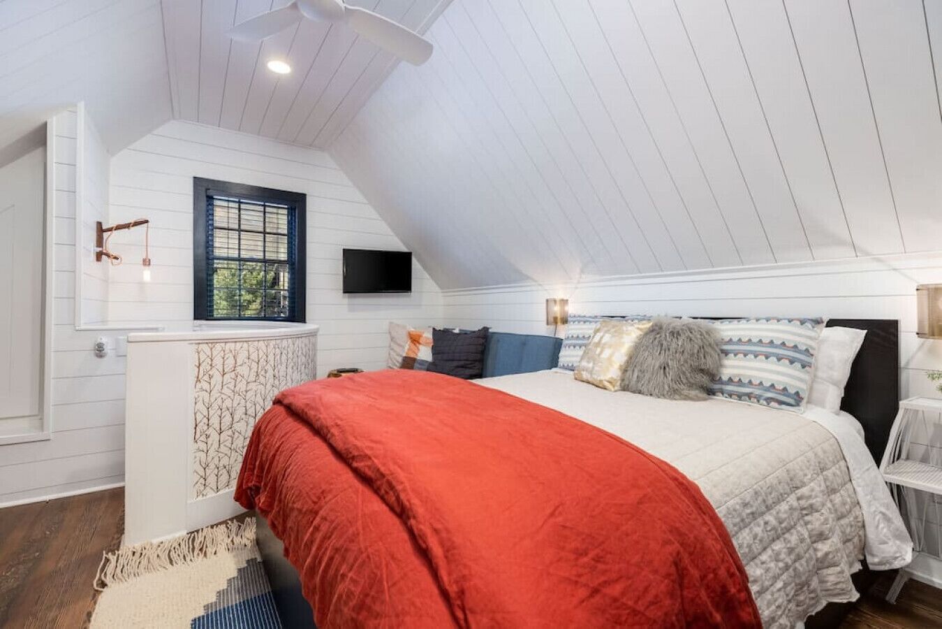 A bedroom with a sloped ceiling, a bed with multiple pillows, a window with blinds, and a wall-mounted TV.